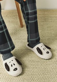 Bílé plyšové pantofle s hnědýma ušima a rysy psa. Nosené s šedými ponožkami, doplněné kostkovanými pyžamovými kalhotami. Viditelná texturovaná podložka na podlaze.