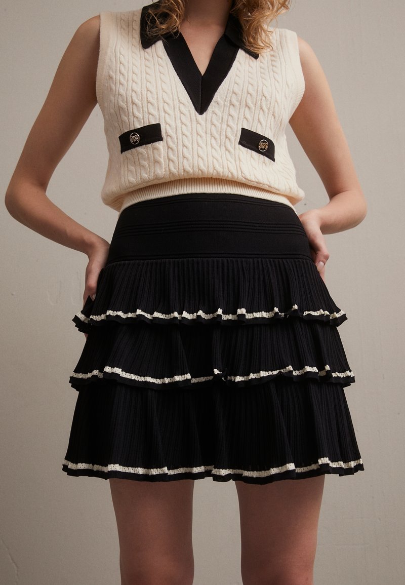 Krémová vestová sarká s pleteným vzorem a černým límcem a akcenty, spárovaná s černou skládací sukní se třemi volánovými vrstvami a bílým lemem.