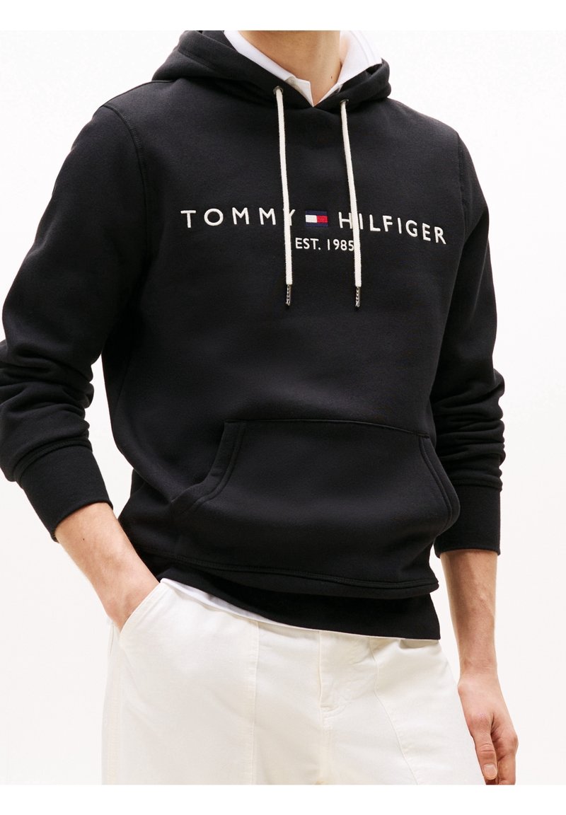 Sudadera negra hecha de tela suave. Cuenta con cordones blancos y un bolsillo frontal. Muestra el logo "TOMMY HILFIGER EST. 1985".