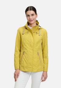 Gelbe wasserdichte Jacke mit Reißverschlusstaschen, Kordelzugkapuze und vertikalen Nähten für einen taillierten Look. Leichtes Material.