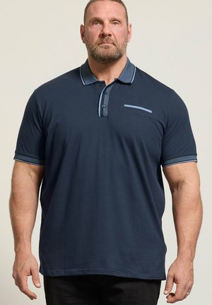 Muskeløs mand iført en mørkeblå poloshirt med korte ærmer og lyseblå trim på krave, stolpelukning, ærmekanter og brystlommedetalje.