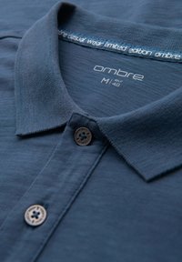 Mörkblå pikétröja med texturerad tyg, med krage och en knapp. Etiketten lyder "ombre casual wear limited edition."