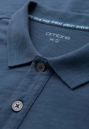 Polo de color azul oscuro con una tela texturizada, que presenta un collar y un botón. La etiqueta dice "ombre casual wear edición limitada."