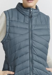 Gilet matelassé bleu avec un col haut et une fermeture éclair à l'avant. Fabriqué en matériau léger avec un design texturé et gonflé.