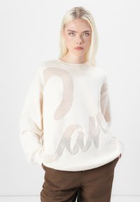 Krämfärgad oversized stickad tröja med en texturerad design och utskurna bokstäver. Har en rund halsringning och ribbade ärmslut, samt matchas med bruna byxor.