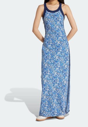 Femme portant une robe maxi sans manches à fleurs bleues avec bordure marine et baskets blanches, debout les mains derrière le dos.