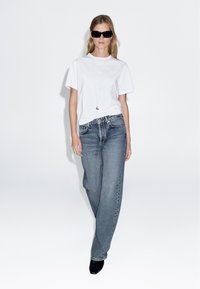 Weißes T-Shirt mit kurzen Ärmeln kombiniert mit lockeren grauen Jeans. Das Outfit wird durch schwarze spitze Stiefeletten und dunkle Sonnenbrillen ergänzt.