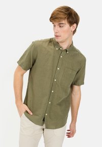 Korte mouwen overhemd in olijfgroene katoen, met een borstzak en subtiele textuur. Model poseert met hand in de zak.