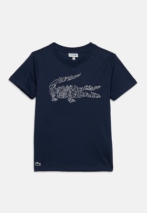 Tumesinine lühikeste varrukatega T-särk valge kursiivkirjaga, mis kujutab rinnal krokodilli kujutist, ja väike roheline krokodillilogoga äärisel.