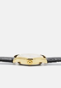 Boîtier de montre en or avec un cadran rond, bracelet en cuir noir, surface lisse et une couronne sur le côté. Design minimaliste classique.