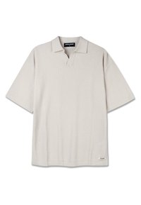 Camisa de polo de punto beige de manga corta con patrón texturizado, cuello abierto y pequeña etiqueta de la marca en el dobladillo inferior.