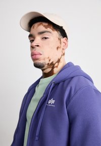 Fialová mikina s kapucňou na zips a logom, nosená cez svetlozelenú košeľu. Taktiež obsahuje béžovú baseballovú čiapku. Hladká textúra, športový dizajn.