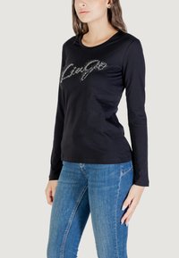 Modella che indossa una maglietta nera a maniche lunghe con testo luccicante "Lingo", abbinata a jeans blu, su uno sfondo neutro.