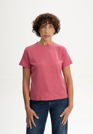 Vrouw met kort krullend haar, gekleed in een effen roze shirt met korte mouwen en een blauwe spijkerbroek, staande tegen een witte achtergrond.
