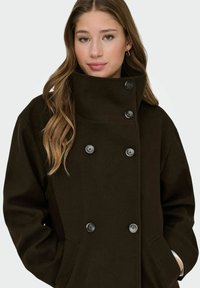 Chaqueta marrón de doble botonadura con cuello alto, que presenta grandes botones negros y bolsillos laterales. Acabado de tejido suave y texturizado.
