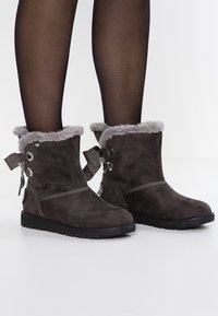 Botines de ante gris con ribete de piel sintética, lazo decorativo y suela de goma; cuenta con botones laterales y un diseño de punta redonda.