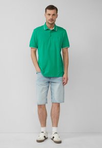 Zielona koszulka polo z białymi akcentami przy kołnierzyku, połączona z jasnoniebieskimi szortami dżinsowymi i białymi trampkami. Prosty fason z krótkimi rękawami.