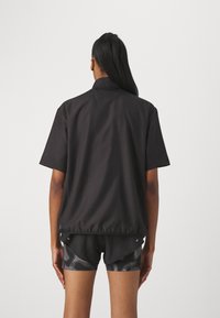 Veste noire à manches courtes avec col, présentant une coupe ample et un ourlet incurvé. Portée avec un short à motifs, le tissu semble lisse.