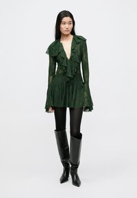 Zelené průsvitné šaty s volánovým límcem a manžetami, s vrstveným designem a květinovým vzorem; doplněné černými kozačkami ke kolenům a punčochami.