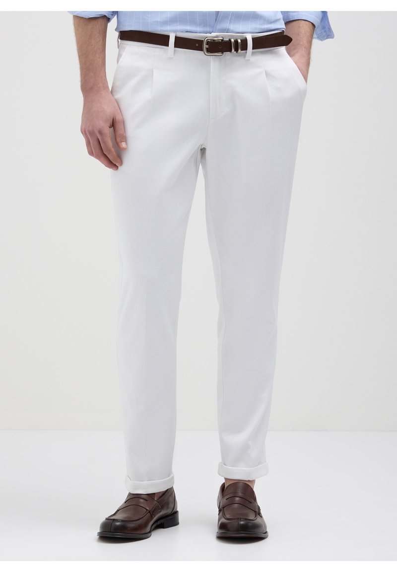 Homme portant un pantalon blanc sur mesure avec revers roulés, ceinture en cuir marron et mocassins en cuir marron, debout avec les mains partiellement dans les poches.
