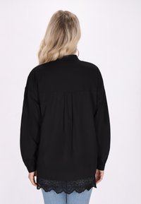 Blusa nera con orlo in pizzo, maniche lunghe e colletto con bottoni. Il tessuto appare morbido con un fit rilassato e un dettaglio plissettato sul retro.