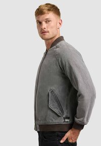 Homme aux cheveux blonds courts portant une veste aviateur grise zippée avec poignets et ourlet côtelés, vu de profil sur un fond gris.