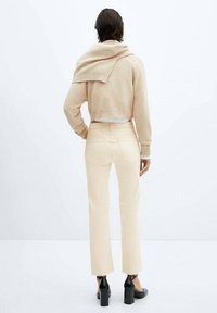 Pull en laine beige drapé sur les épaules, associé à un jean taille haute de couleur claire et des chaussures à talons blocs noires. Fond uni.