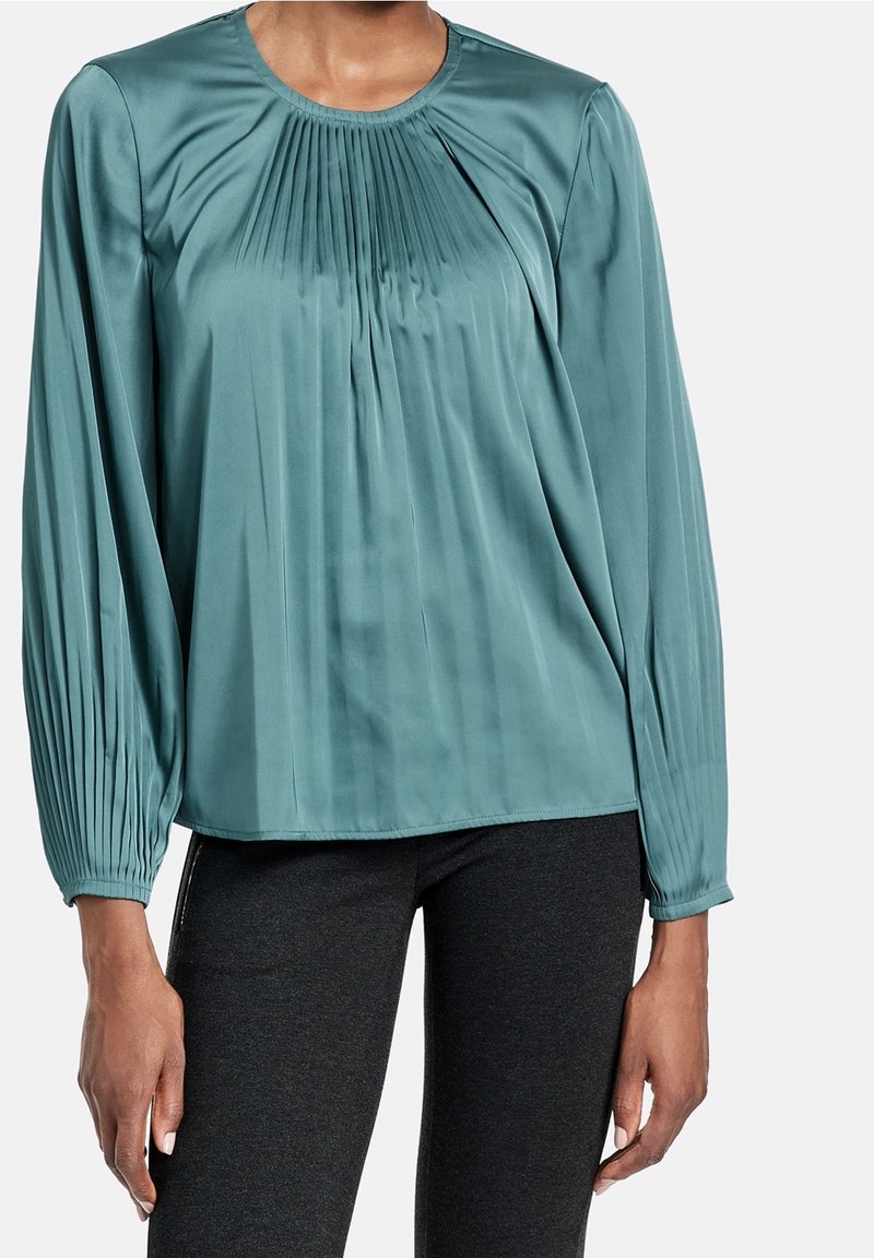 Blusa di satin turchese con maniche lunghe, design frontale plissettato, scollatura rotonda e dettagli arricciati sulle spalle. Indossata con leggings scuri.