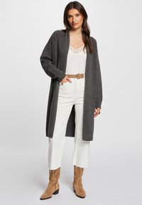 Mörkgrå lång cardigan med vida ärmar, lager över en vit topp med spetsdetaljer. Matchad med högmidjade vita jeans och bruna ankelboots.