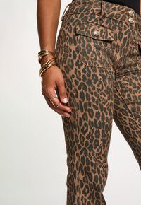 De hand van een vrouw met gouden armbanden en ring rustend nabij een broek met luipaardprint, met knoopzak en details van de tailleband.