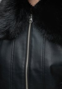 Gros plan d'une veste en cuir noire avec une fermeture éclair argentée et un large col en fourrure noire.