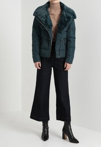 En teal puffjacka med krage i fuskläder, dragkedjestängning och sidofickor, tillsammans med vida mörka jeans och spetsiga svarta ankelstövlar.