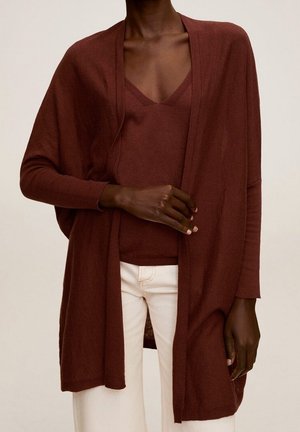 Personne à la peau foncée portant un pull marron col V et un long cardigan assorti, associé à un pantalon blanc, debout sur un fond neutre.