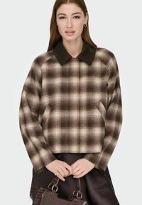 Kvinne iført brun og beige rutete jakke med mørkrand, som holder en mørk brun veske med begge hender foran seg.