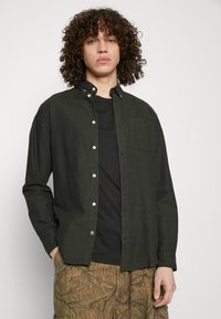 Cămașă butonată verde închis, cu guler, mâneci lungi și un buzunar pe piept, purtată peste un tricou negru, asortată cu pantaloni cu un model.