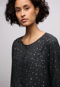 Blusa nera con motivo a pois argentati, scollatura tonda, chiusura con bottoni e maniche lunghe. Tessuto liscio con leggera brillantezza e dettagli arricciati.