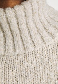 Cremfarbener gestrickter Pullover mit hohem Kragen. Besitzt ein strukturiertes Muster und dezente goldene Fäden, die eingewebt sind. Weiches, voluminöses Material.