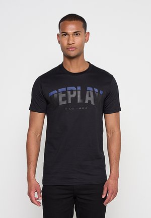 Man stands facing forward wearing a black T-shirt with "REPLAY RIDE HARD" text and black pants against a plain gray background.
