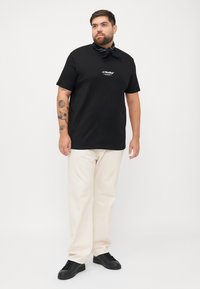 Schwarzes T-Shirt mit "JJ Studios" Logo, kombiniert mit cremefarbenen Hose. Das Modell trägt ein schwarzes Bandana und schwarze Sneakers.