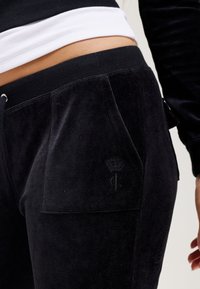 Black velvet joggers with side pockets, featuring a subtle logo and textured fabric. High elastic waistband for a secure fit.