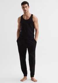 Jeune homme debout pieds nus, portant un débardeur noir sans manches et un pantalon de jogging noir, sur un fond clair uni.
