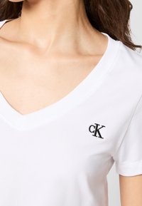Bílý bavlněný tričko s výstřihem ve tvaru V. Na levé straně je černě vyšitá značka "cK". Hladký materiál a krátké rukávy.