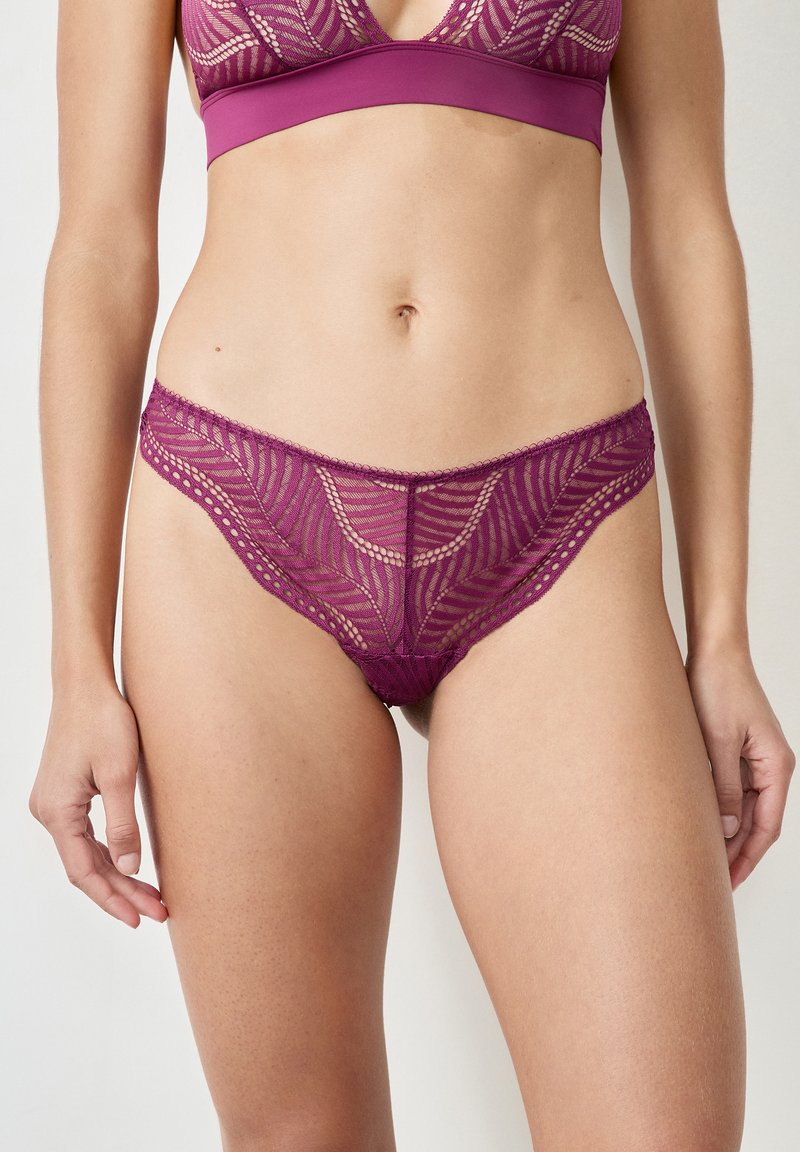 String violet en dentelle avec un motif floral, des bords festonnés et une texture transparente. Le design comprend des sous-couches et des découpes pour le détail.