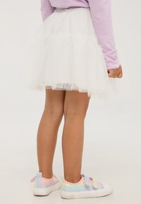 Gonna di tulle bianca con strati arricciati e luccichio, indossata con sneakers pastello arcobaleno. L'elastico in vita è arricciato.