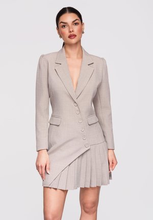 Femme aux cheveux foncés et boucles d'oreilles dorées, portant une robe blazer beige avec des boutons et une jupe plissée, debout devant un fond uni.