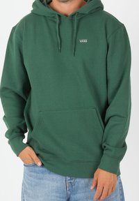Sudadera con capucha verde hecha de tela suave con un bolsillo frontal y capucha con cordón. Presenta un pequeño logo "VANS" en la parte superior izquierda del pecho.
