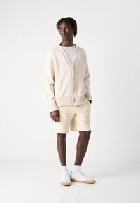 Cream cardigan with ribbed cuffs and a V-neck, paired with light yellow textured shorts and white sneakers with beige accents.