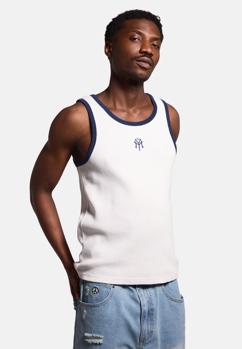 Weißes geripptes Tanktop mit marineblauen Akzenten und Logo auf der Brust, kombiniert mit hellblauen, verwaschenen Denim-Shorts.