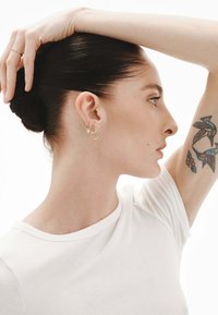 Pendientes de aro de oro con diseños y charms interconectados, llevados por una mujer con un tatuaje, sobre un fondo blanco liso.
