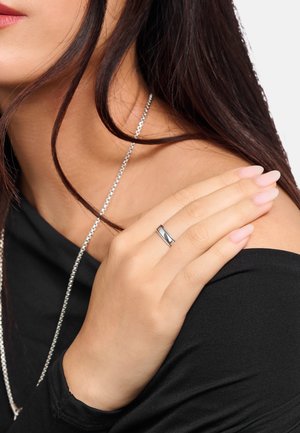 Frau, die einen silbernen Ring am Ringfinger und eine silberne Kettenhalskette trägt, mit dunklen Haaren und schwarzem schulterfreiem Oberteil.